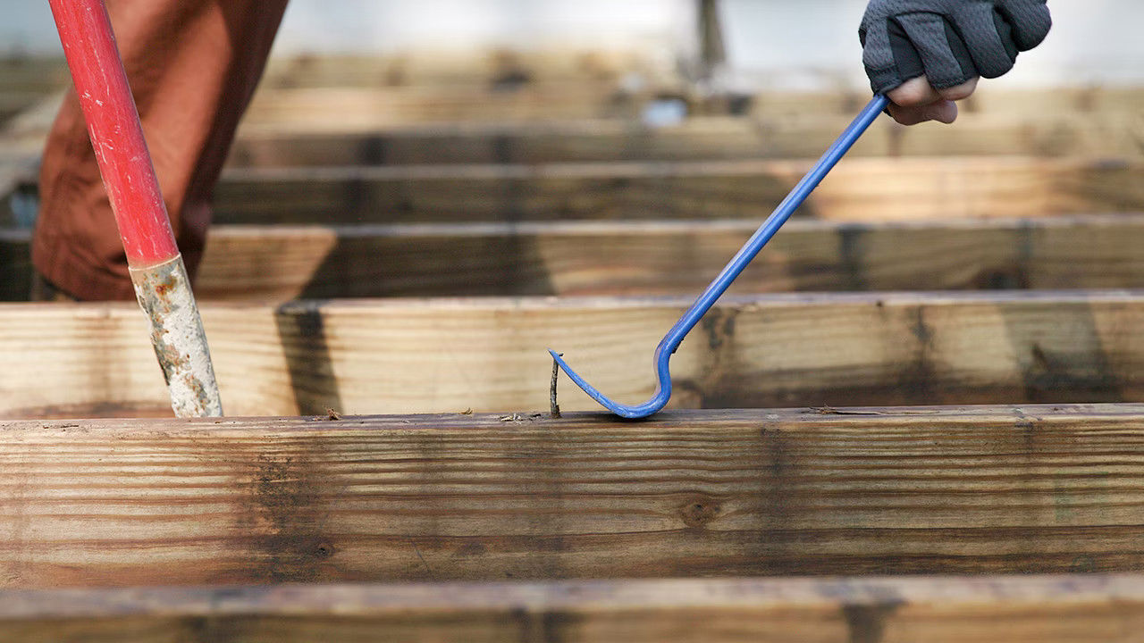 A person uses a blue crowbar to remove nails from wooden planks. The focus is on the tool and wood, conveying a theme of construction or renovation.