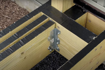 Wooden deck framing with three joists connected by metal brackets. Black decking tape covers the top edges for protection. Background shows mulch and building siding.