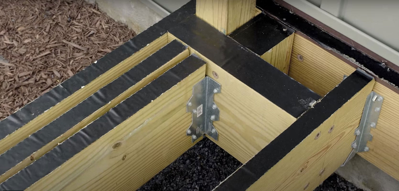 Wooden deck framing with three joists connected by metal brackets. Black decking tape covers the top edges for protection. Background shows mulch and building siding.