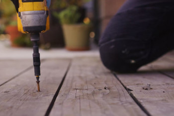 Close-up of a yellow drill driving a screw into a wooden plank, with a blurred background of potted plants and a person kneeling nearby.
