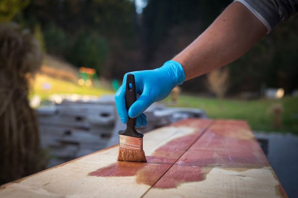 A person with blue gloves uses a brush to apply wood stain on wooden planks outdoors.