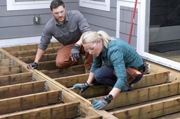 A man and woman wearing safety glasses and gloves measure wooden deck beams with a tape measure. They work attentively next to a house wall.
