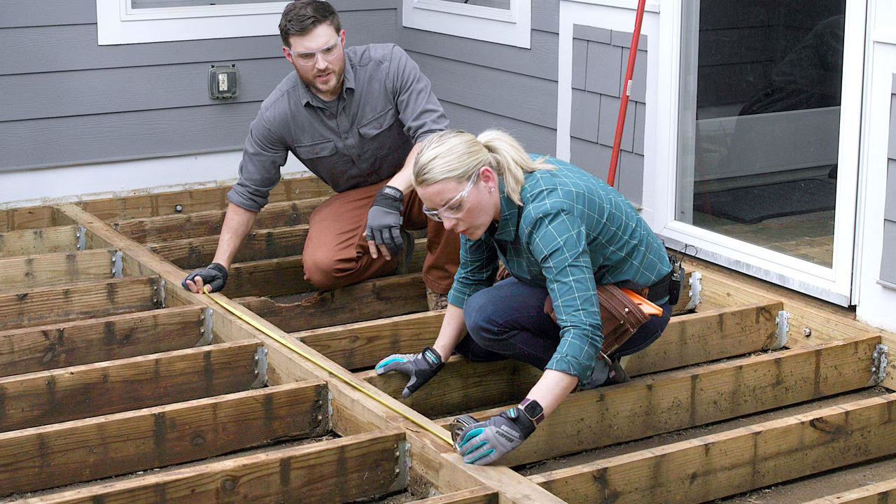 A man and woman wearing safety glasses and gloves measure wooden deck beams with a tape measure. They work attentively next to a house wall.